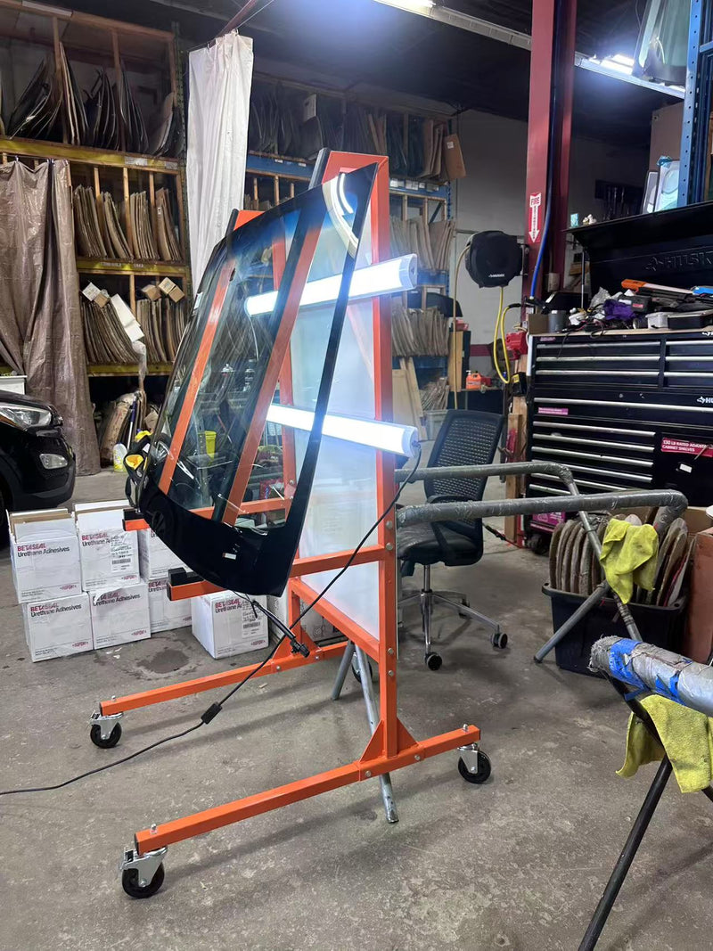Car windshield on an orange stand in a workshop setting with tools and equipment around.